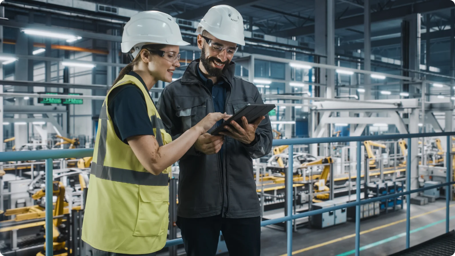 A man and a woman dressed in high visibility vests, using a tablet. It looks like they are in a manufacturing facility. 